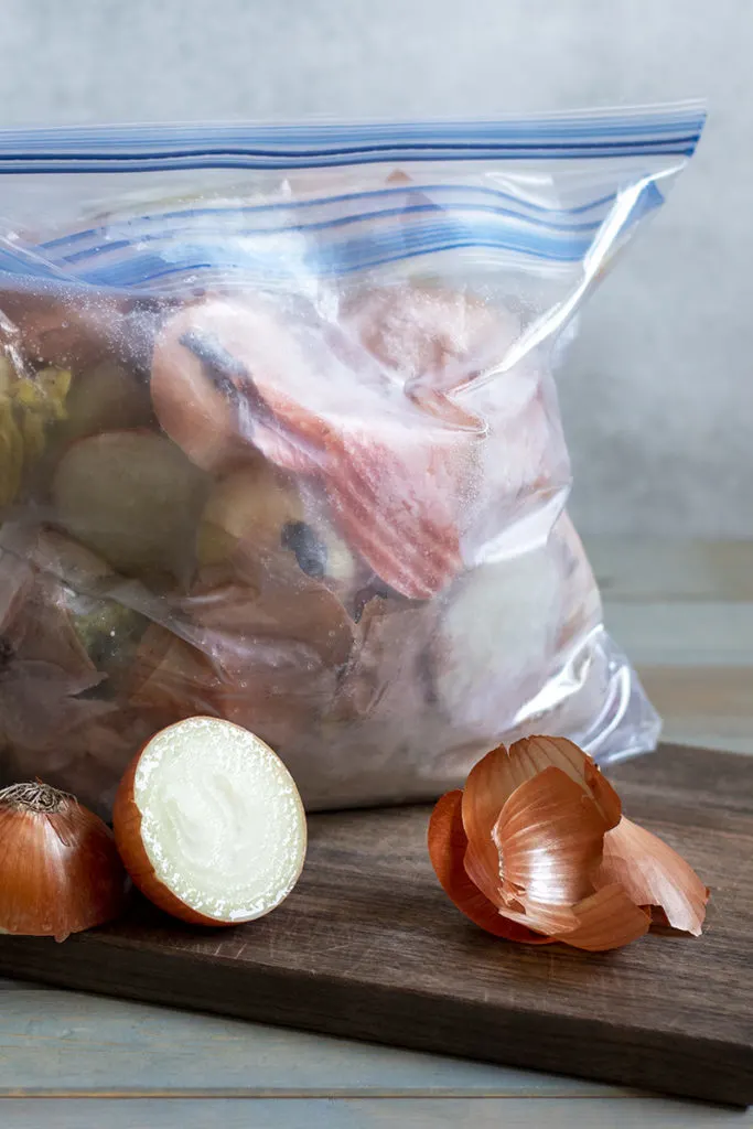 A freezer bag full of kitchen scraps within another freezer bag is sitting on a cutting board next to an onion sliced in half.