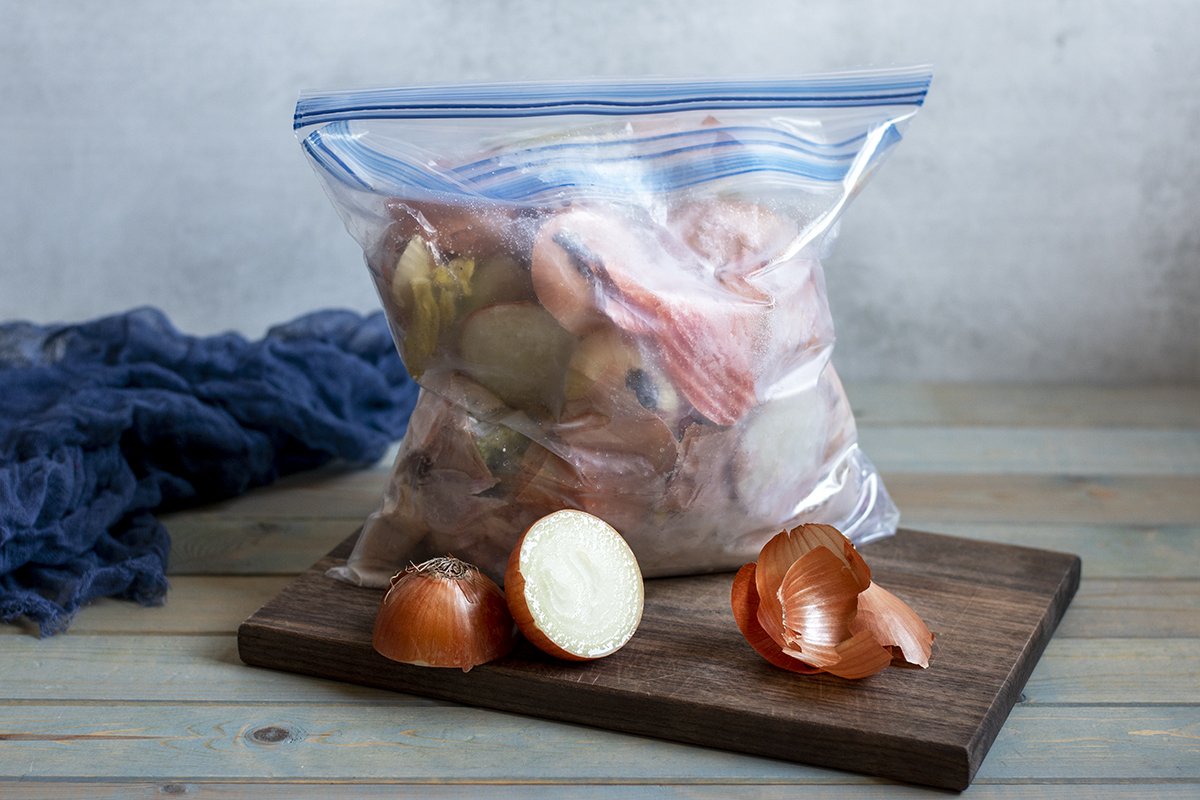 A freezer bag full of kitchen scraps within another freezer bag is sitting on a cutting board next to an onion sliced in half. A blue cloth is decoratively placed in the background.