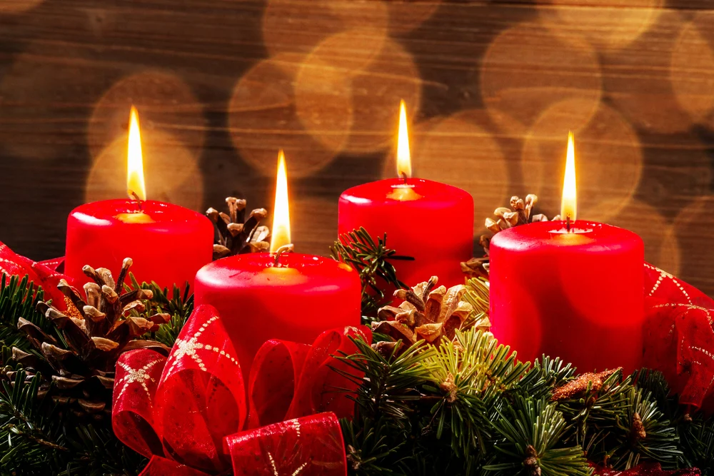 Four burning red pillar candles in a Christmas centerpiece with pine cones, bows, and evergreens.
