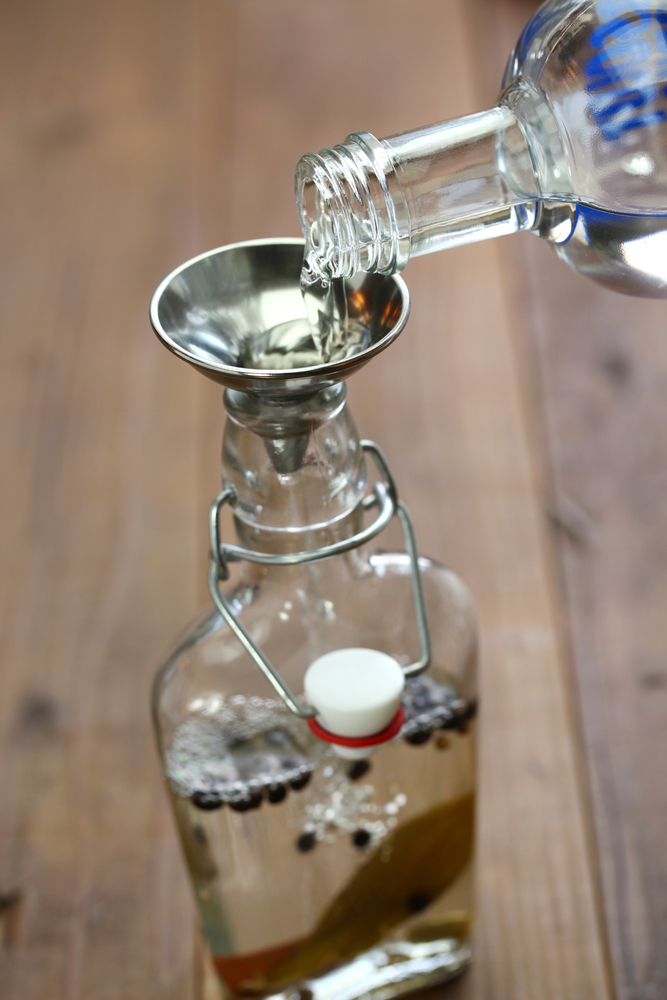 A bottle being filled with vodka to make homemade gin, there are herbs in the bottle. 
