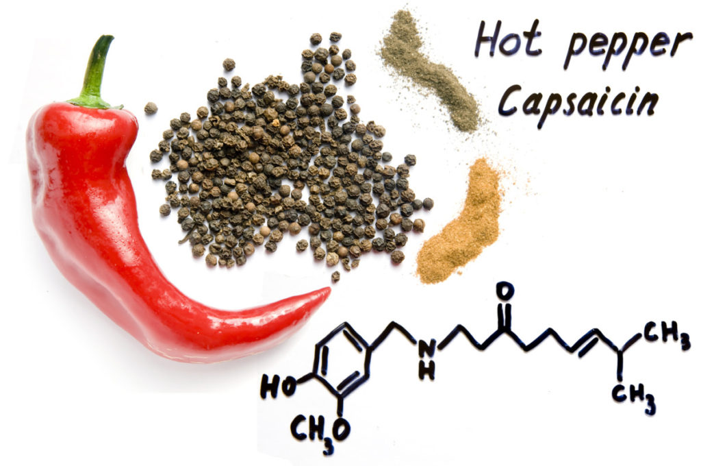A red hot pepper rests on a white background next to various pepper powders. The chemical formula for capsaicin is written out in black marker. 