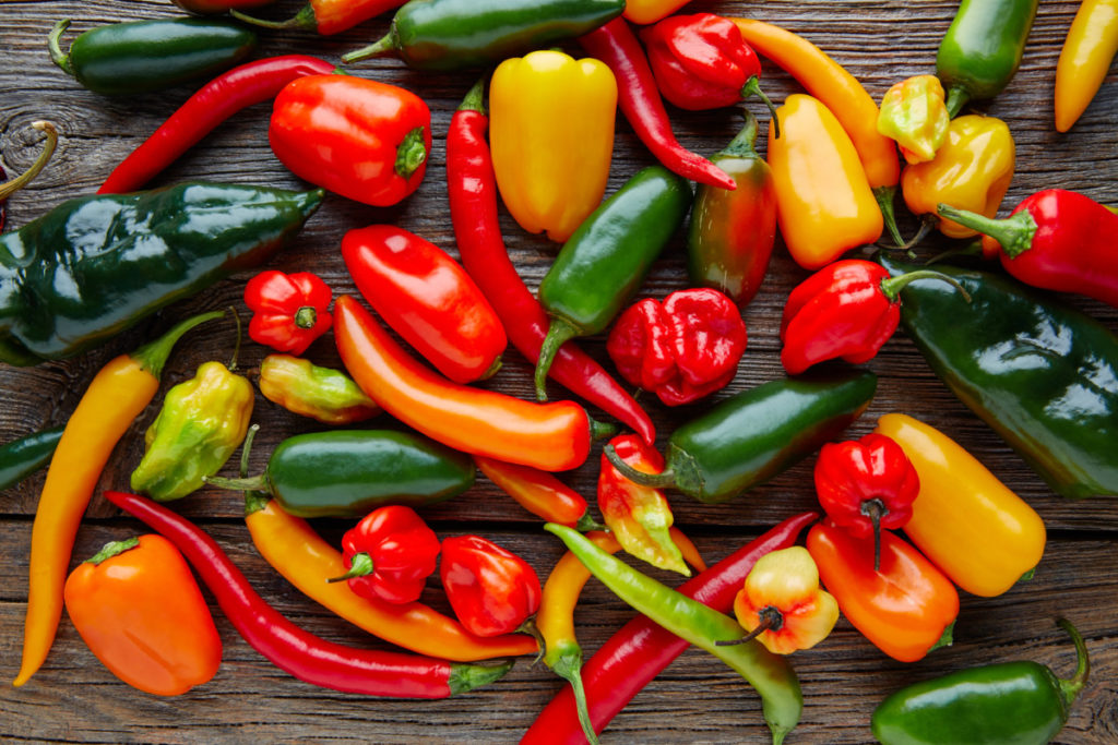 Green, red, orange and yellow hot peppers are strewn across a wooden table top. 