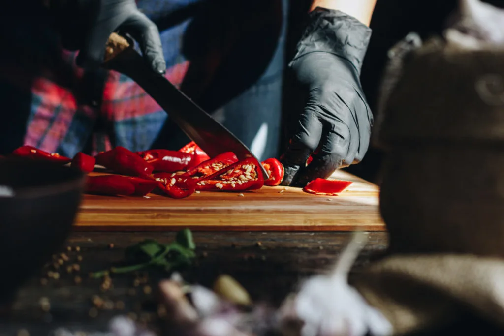 A man wearing black nitrile gloves slices hot peppers on a cutting board.