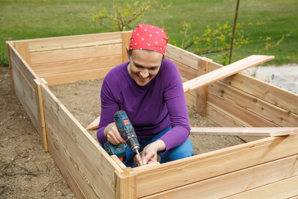 A smiling woman uses a power drill to put together a raised bed.