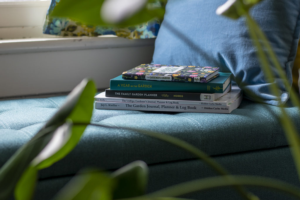 A stack of garden planning books sitting on a bench under a window, there is a plant in the foreground.