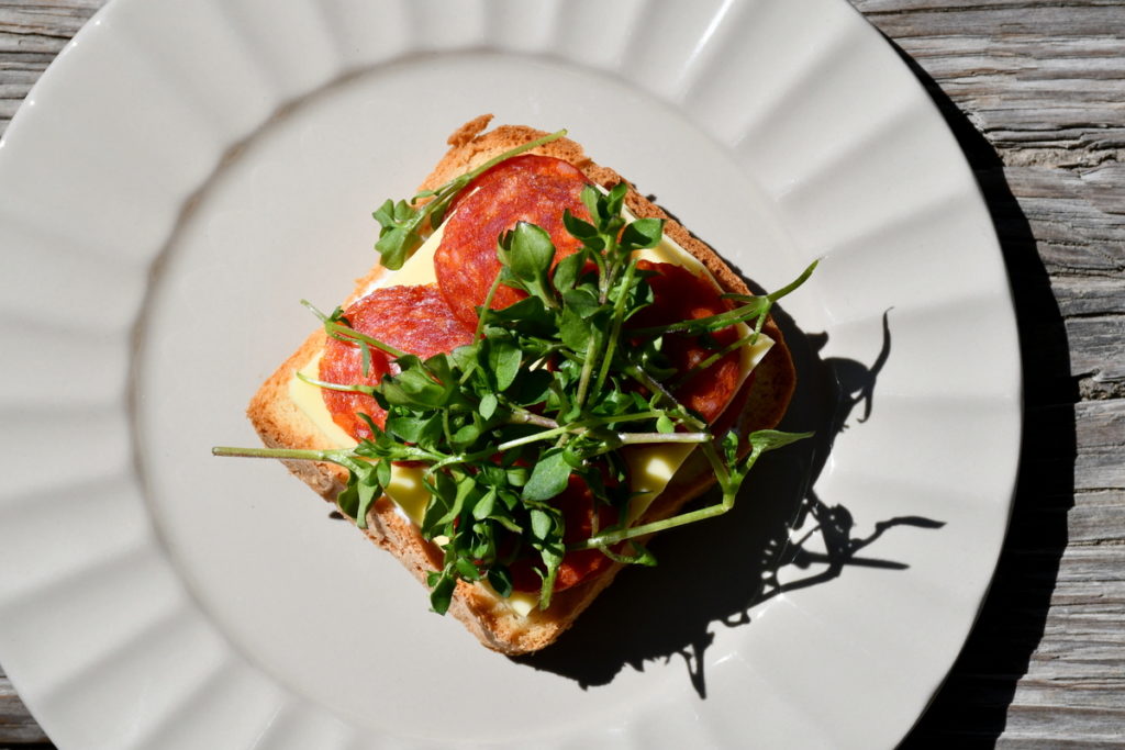 A white plate with a piece of toast with cheese, pepperoni and chickweed on it. 
