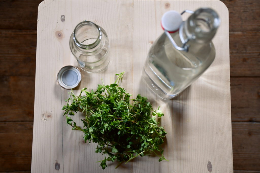 Aerial view of a bottle of vodka, an empty jar and lid, and a pile of chopped chickweed stems. All are on top of a light colored wood cutting board.
