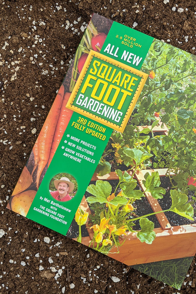 A copy of the newest edition of Square Foot Gardening laying in the soil. 