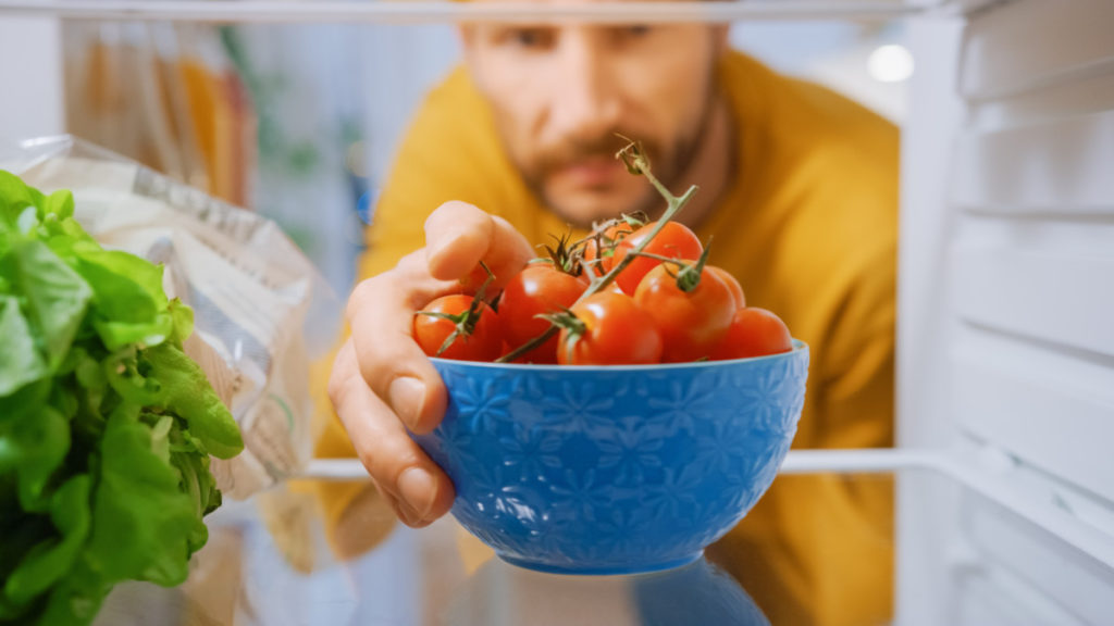 A man reaching for a bowl of tomatoes in a fridge.