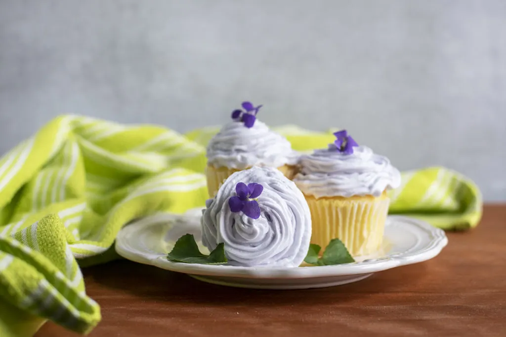 Cupcakes with violet buttercream frosting on a plate. A bright green tea towel is in the background.