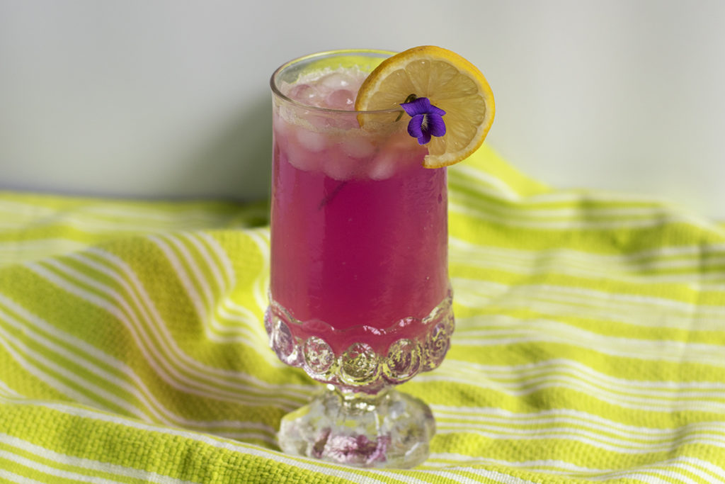A glass of hot pink violet lemonade with a lemon and violet garnish.