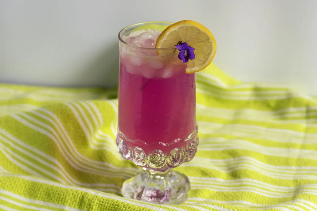 A glass of hot pink violet lemonade with a lemon and violet garnish.