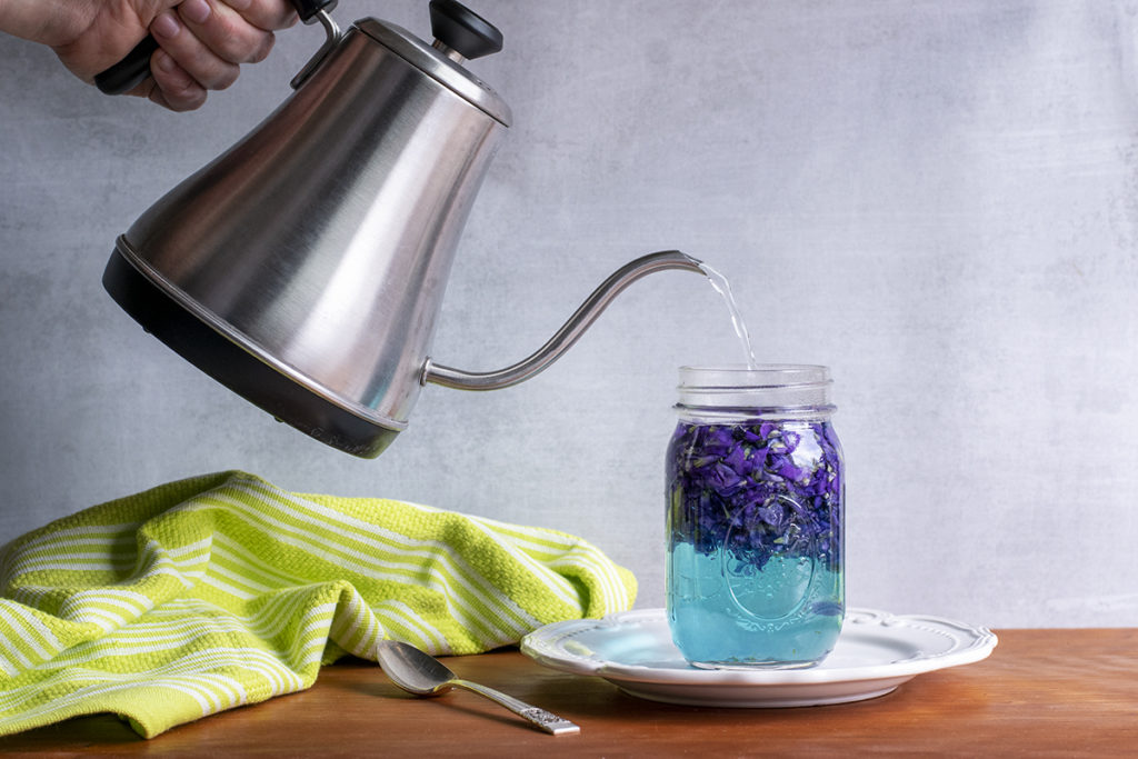 A hand is shown holding a tea kettle, pouring water into a jar of violet petals. The water in the jar is turning blue.