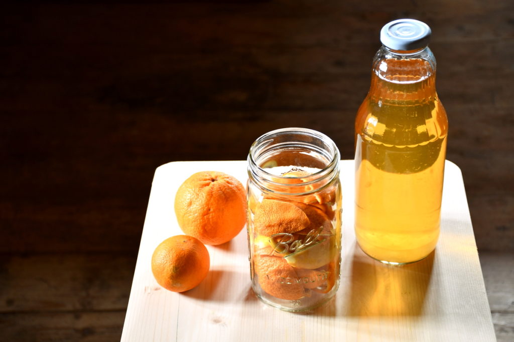 A plain pine table top with a the jar of apple scrap vinegar, a mason jar filled with orange scraps and a two oranges on the table next to it.
