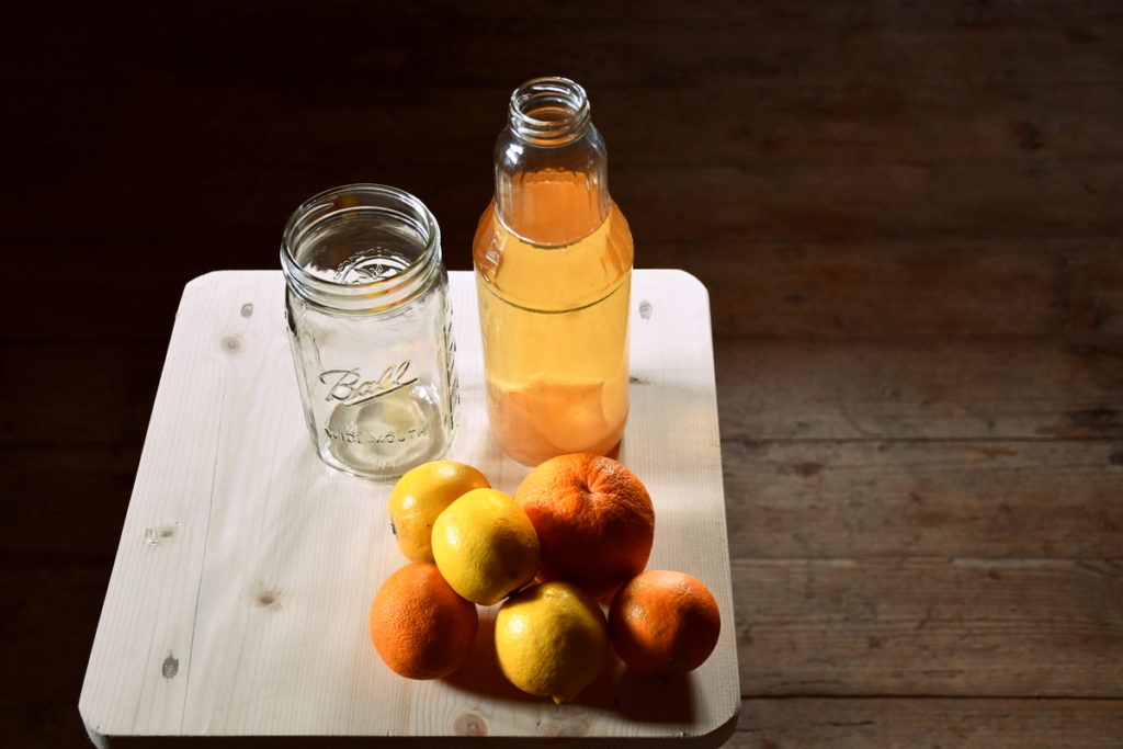 A small wood table with an empty jar, a pile of citrus fruits and a jar of apple scrap vinegar.