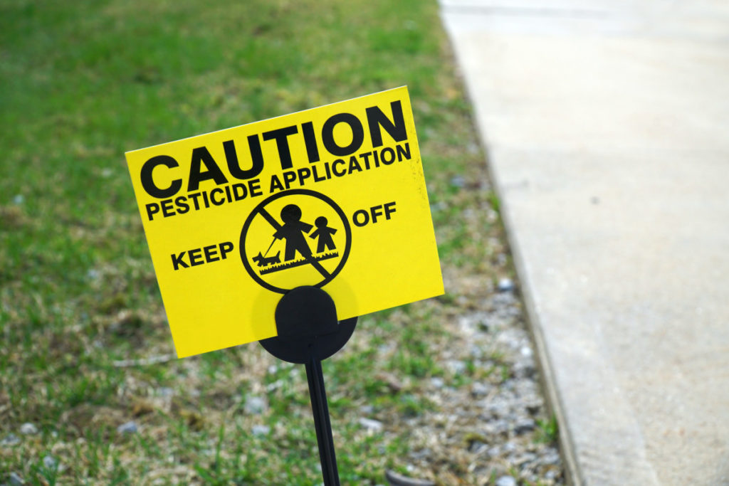 A pesticide application sign stuck in the edge of a lawn. It reads, "Caution pesticide application keep off"