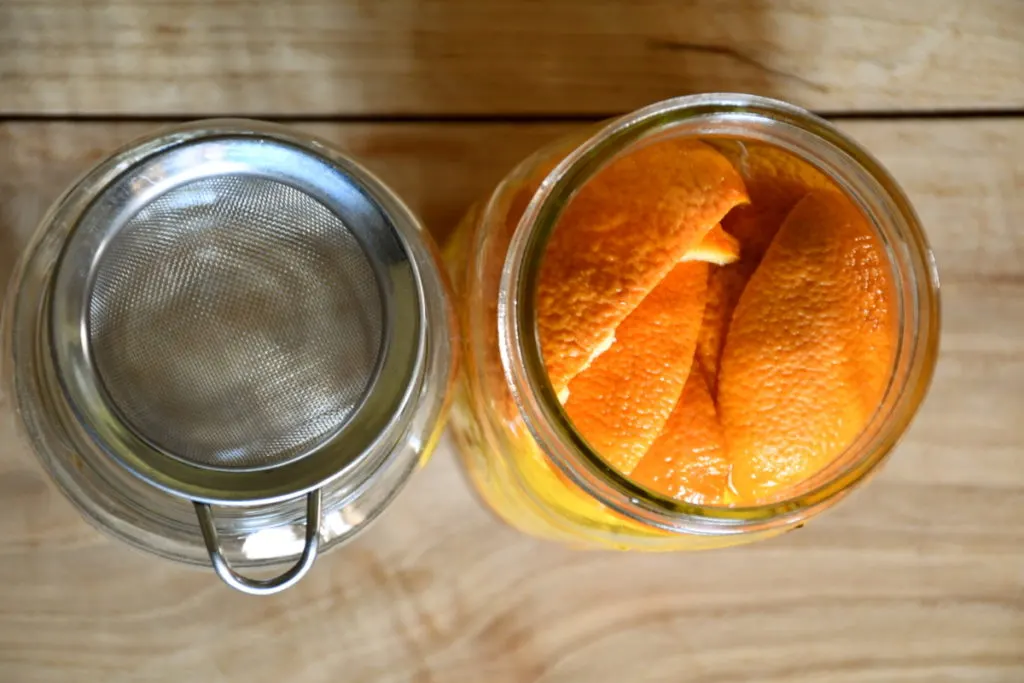 Overhead view of two jars. The jar on the left is empty and has a tea strainer over the mouth. The jar on the right is filled with vinegar and orange peels.