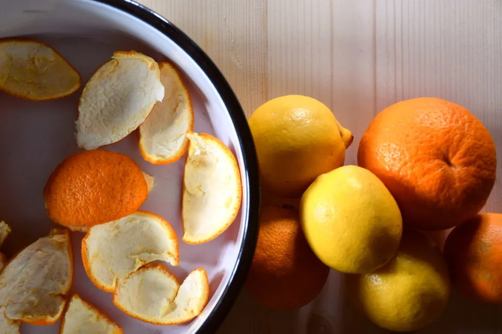 An enamel bowl with orange peels in it sits on a table top. To the right of the bowl is an assortment of oranges and lemons.