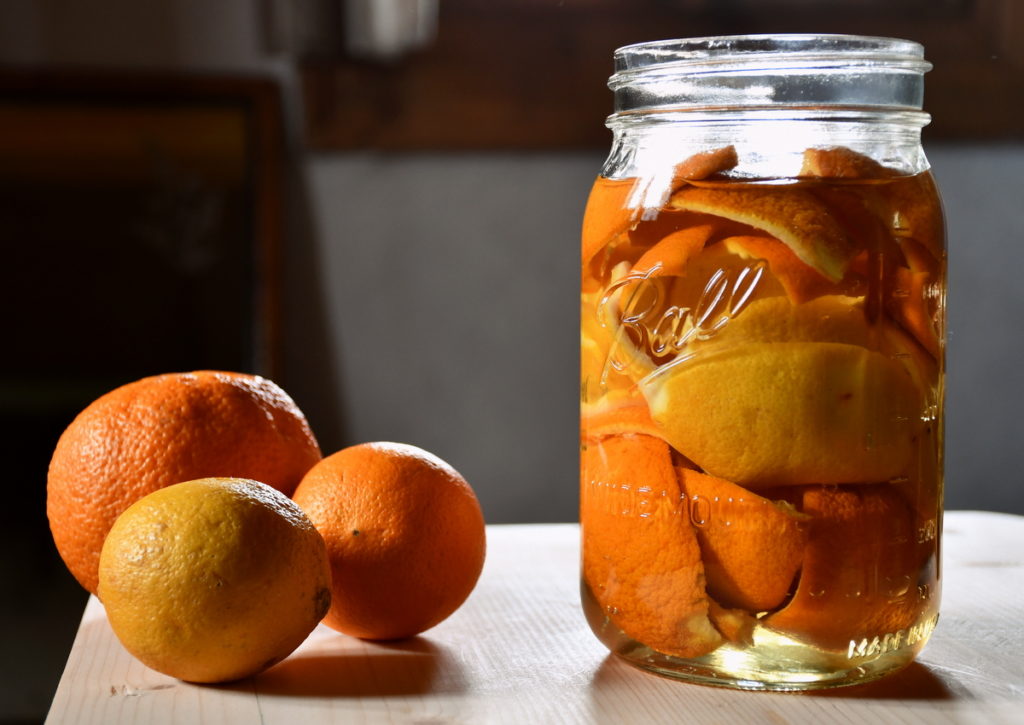 On the right is a mason jar filled with vinegar and orange peels. On the left is an orange, a clementine, and a lemon. All are sitting on a table. 