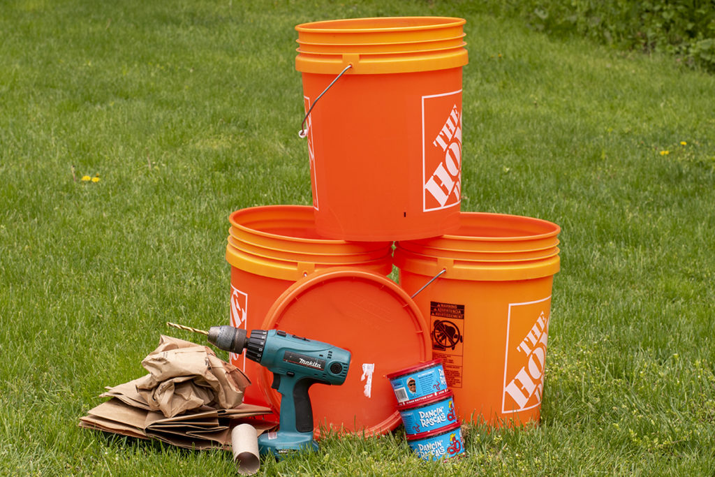 Three 5-gallon buckets, a lid, three tubs of worms, an electric drill and a pile of paper bags are all stacked together on a lawn.