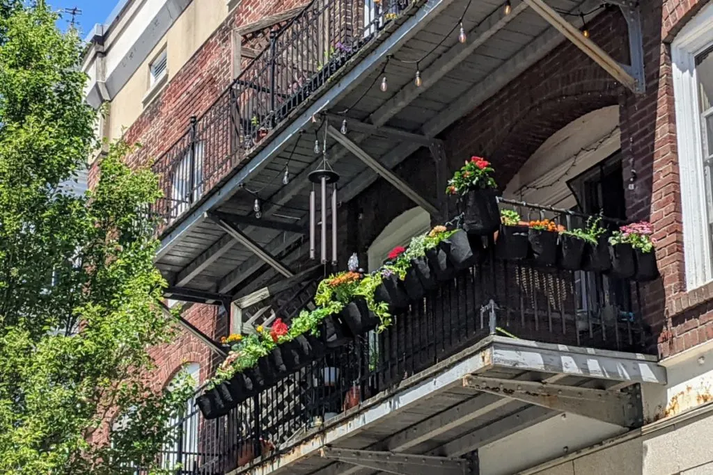 A couple dozen black, cloth grow bags hang from a balcony railing outside. Flowers and herbs are growing in the grow bags. There is a windchime hanging from the ceiling of the balcony as well as outdoor lights. The building is a brick building, there is a tree on the left of the balcony. The sun is shining. 