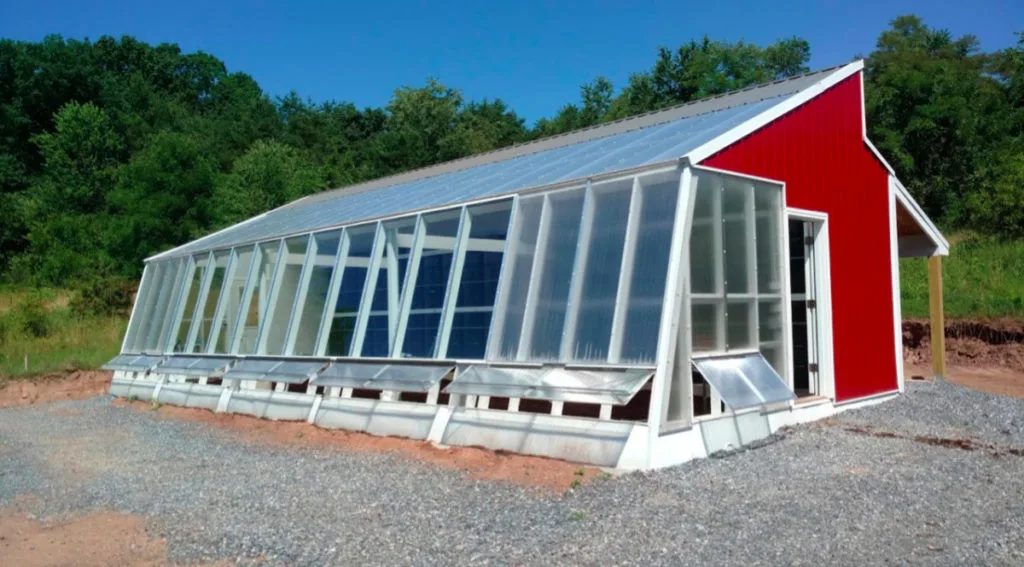 The exterior of a sustainable greenhouse with glass windows, open vents, red metal siding, and polycarbonate sheets for part of the roof.