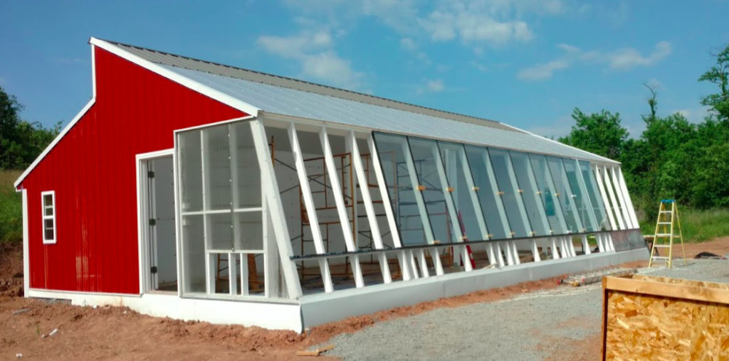 A view of the south wall of the sustainable greenhouse with most of the glass window panels installed. The red metal siding on the west gable end is visible.