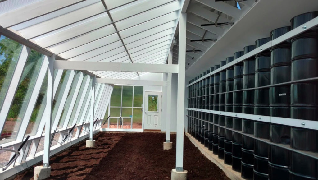 The sunny interior of an experimental sustainable greenhouse with white framing, glass windows, mushroom soil, and a wall of black steel barrels.