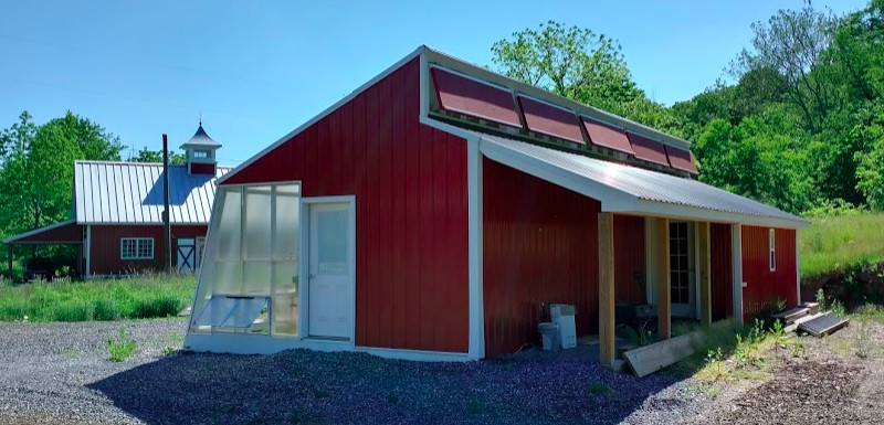 A view of the rear of the sustainable greenhouse showing a rear porch with brown wood columns, the experimental vents near the ridge of the roof, which are red, and red metal siding on the back of the greenhouse and on the east gable end.
