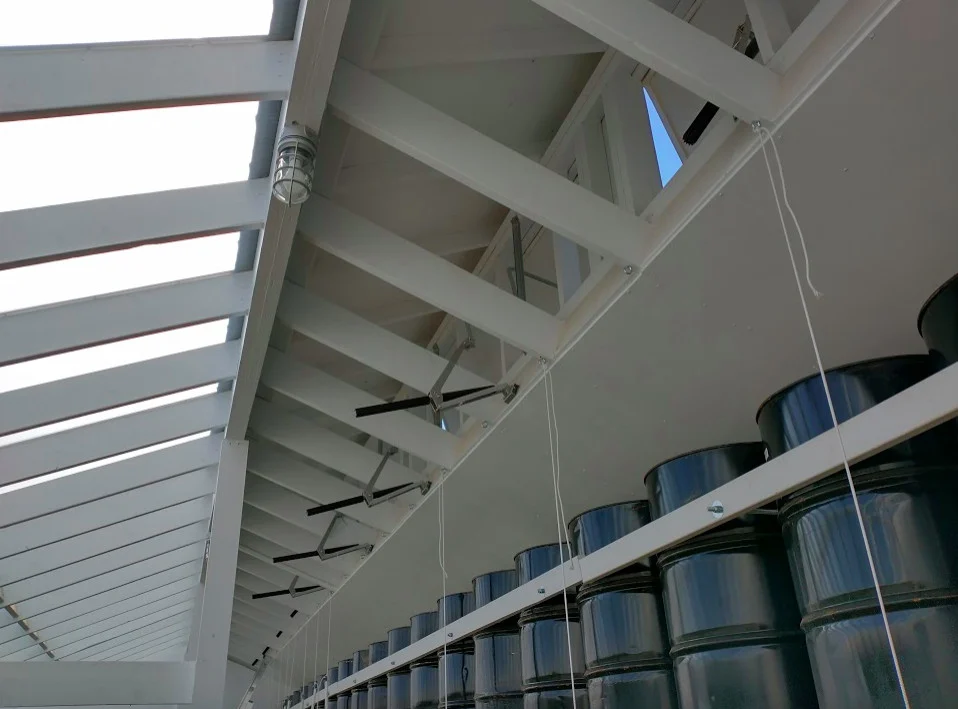 A view of the white-framed ceiling of the sustainable greenhouse with a row of experimental vent openers and a row of black steel barrels visible.