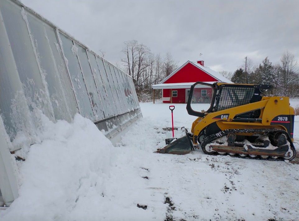 A winter scene in front of the sustainable greenhouse with a huge snow drift. A yellow Caterpillar skid loader has cleared away snow along most of the south wall of the greenhouse. There is a bright red pole barn in the background.
