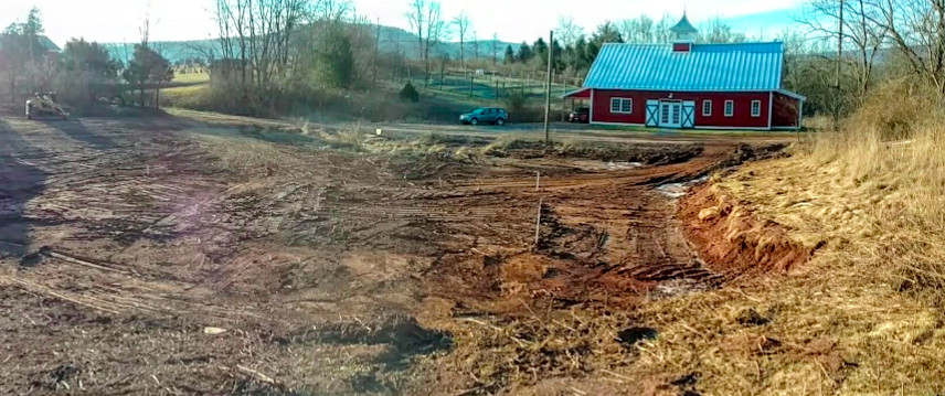 The cleared site for a greenhouse, somewhat muddy, with a red barn in the background and bare trees.