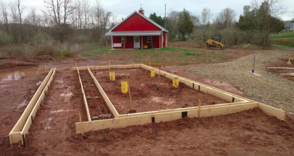 The concrete form work for a sustainable greenhouse, with lumber forms laid out in a square and a red pole barn in the background.