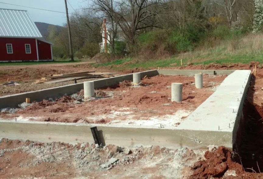 The concrete foundation, recently poured, of a sustainable greenhouse, with bare soil inside, and a red barn in the background.