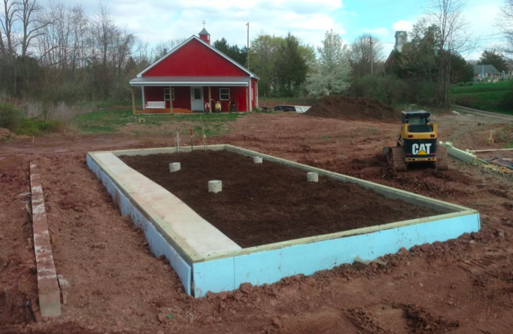 The completed and insulated foundation of a sustainable greenhouse. The insulation is blue. There is a red pole barn in the background, as well as a yellow Caterpillar skid loader.