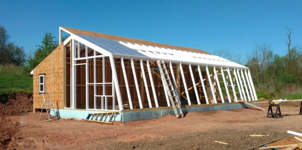 A partially framed sustainable greenhouse. The framing is painted white. A portion of the blue insulation on the foundation is visible. An aluminum ladder leans against the framing.