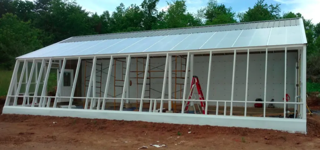 A view of the white framing of the south window wall of the sustainable greenhouse. White polycarbonate panels are visible on the roof, along with gray metal roofing.