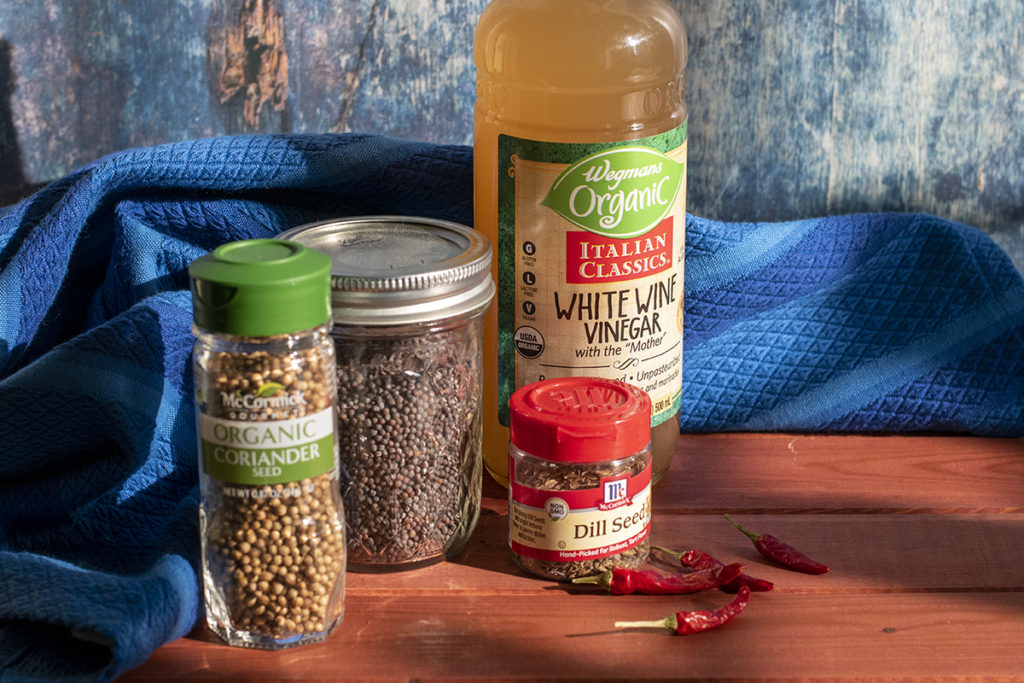 A bottle of Wegmans organic white whine vinegar, McCormick dill seed, McCormick organic coriander seed and a small mason jar of mustard seed placed on a wooden table top. There are four dried chilis next to the jar of dill seed. A royal blue hand towel is draped behind them. 