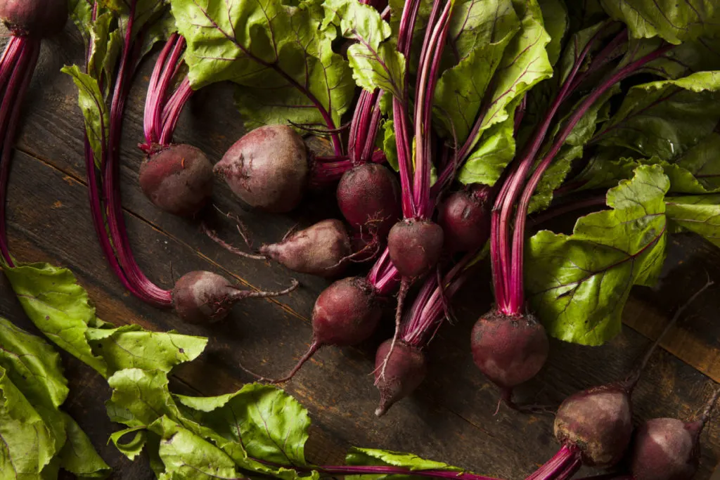 Several red beets with tops are spread across a dark brown, rustic wooden table top. 