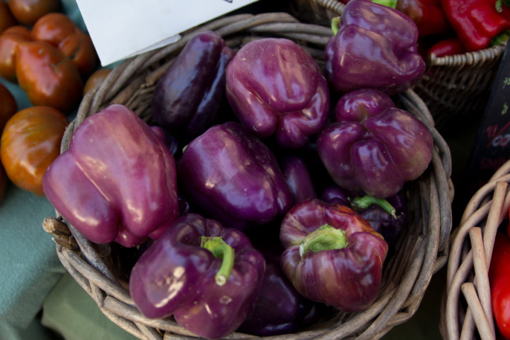 A small woven basket contains rosy, lilac colored bell peppers. There are other baskets of peppers on the periphery. 