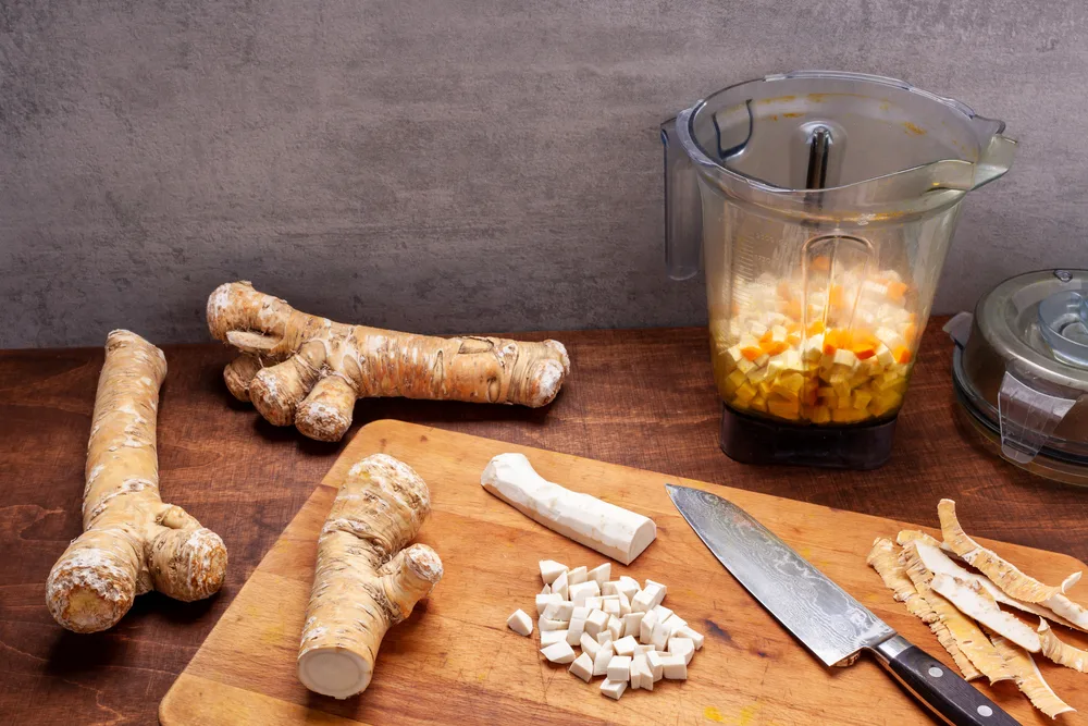  A kitchen counter with a blender cup filled with chopped horseradish pieces. There is also a cutting board with peeled horseradish and the peels. Several unpeeled horseradish roots lay next to the cutting board. There is a Damascus steel chef's knife. on the cutting board. 