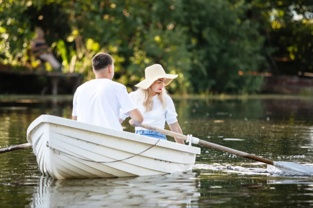 A couple in matching white tshirts are sitting in a rowboat. The man is rowing. The woman is looking into the water. She is wearing a straw sunhat and bright red lipstick. 