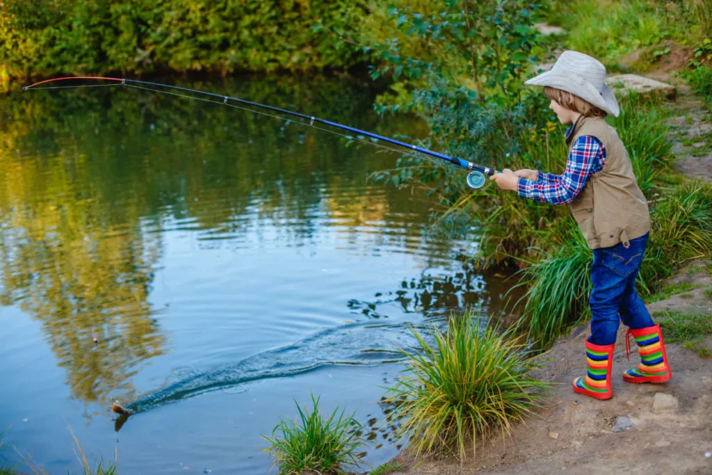 A young boy reals in a fish on a fishing rod. The child is standing next to a small pond. The boy is wearing rainbow striped boots, blue jeans, a  fishing vest, a plaid shirt and a beige sunhat.