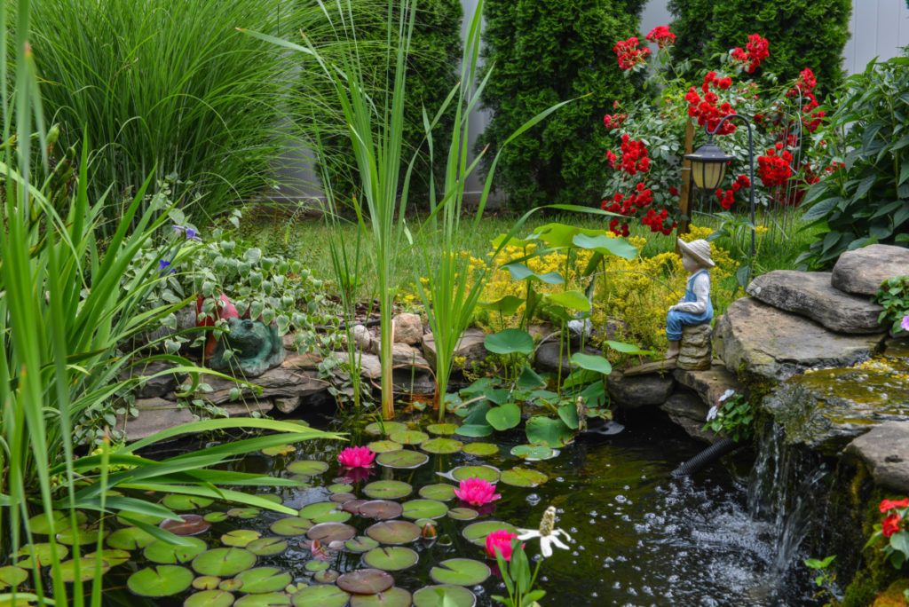 A backyard pond full of water lilies. Small statues decorate the edge of the pond and there are flowers blooming along the edge. There is a stone waterfall on the right.