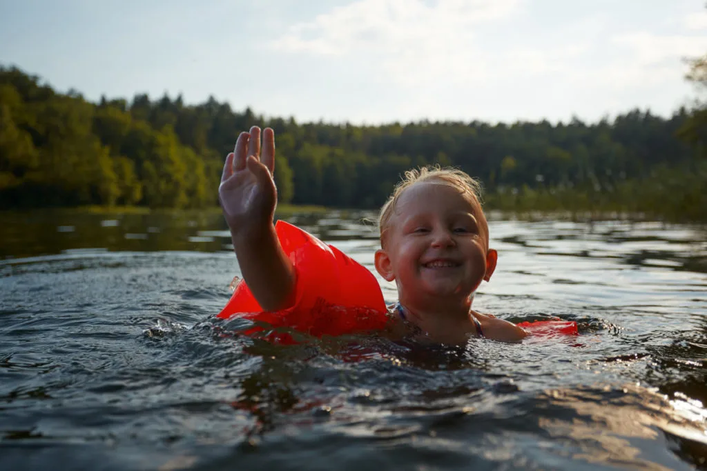 A small child is swimming in a pond, she is wearing bright orange arm floaties and smiling and waving at the camera.