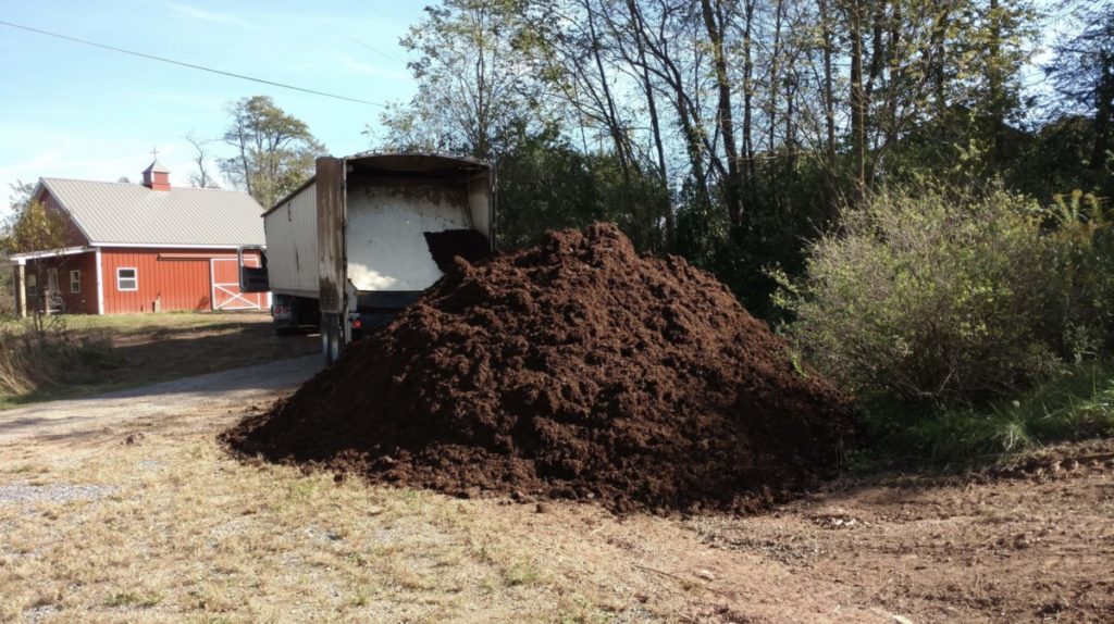 A large mound of brown mushroom substrate sits behind a tractor trailer. The superbarn is in the distance.
