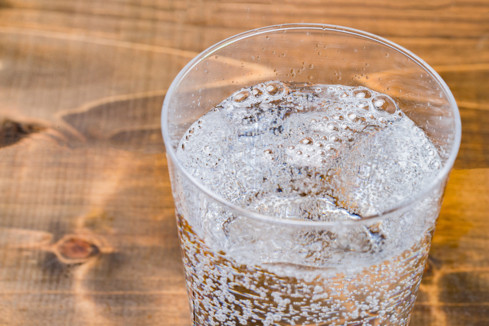 A clear glass contains clear, sparkling soda. The glass is sitting on a wooden table. 