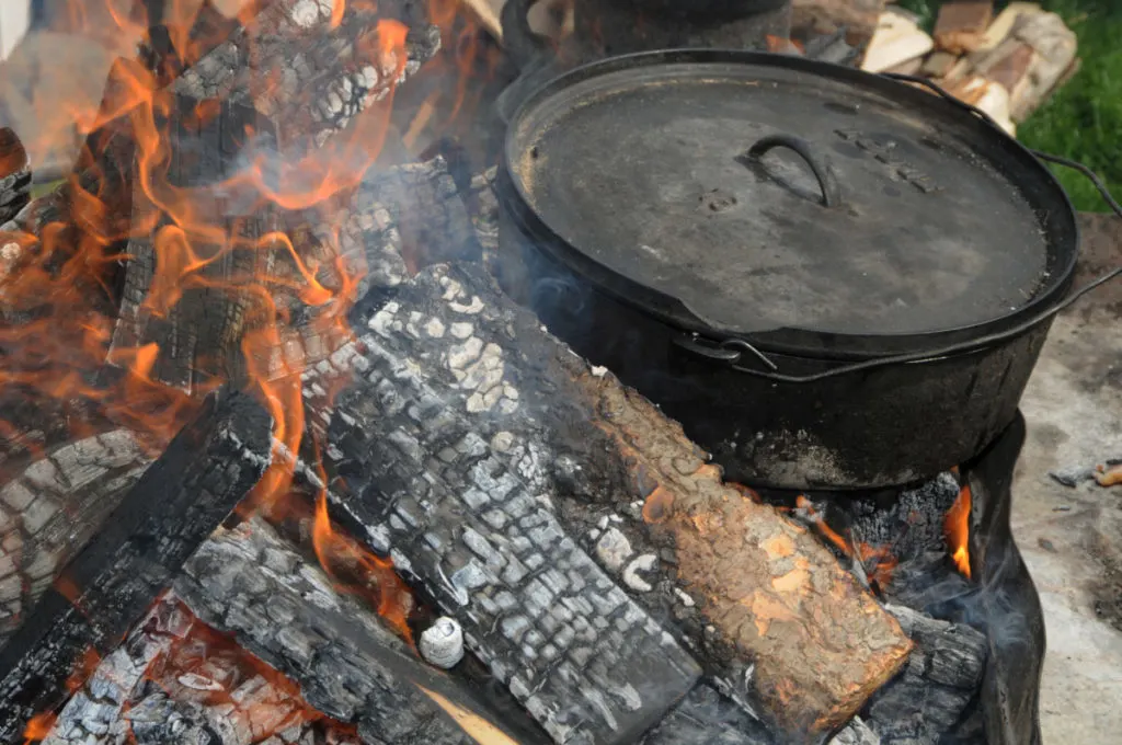 A dutch oven is sitting directly in the coals of a hot fire outdoors.