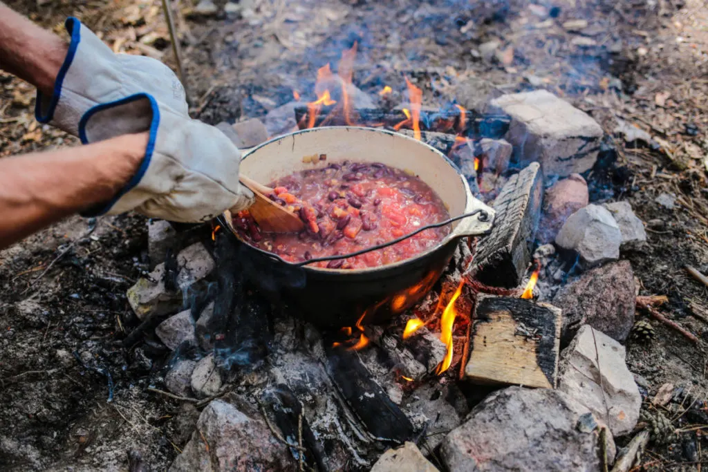 Someone is wearing thick leather gloves and they are holding wood spoons and stirring a pot of chili being cooked over an open campfire.