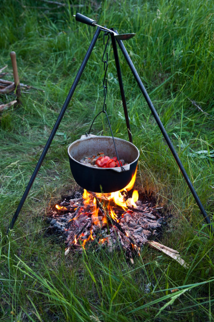 Cast iron tripod holding a dutch oven over a fire. There is food in the dutch oven.